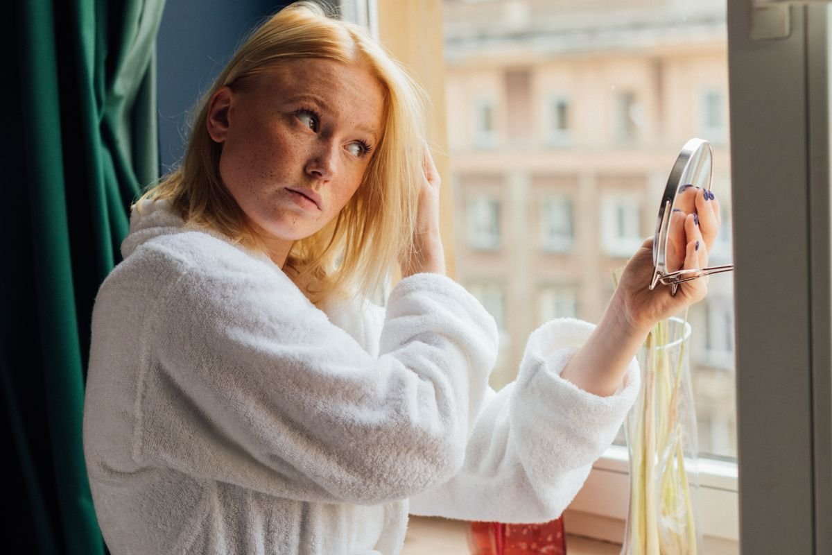 Woman looking into a handheld mirror beside a window