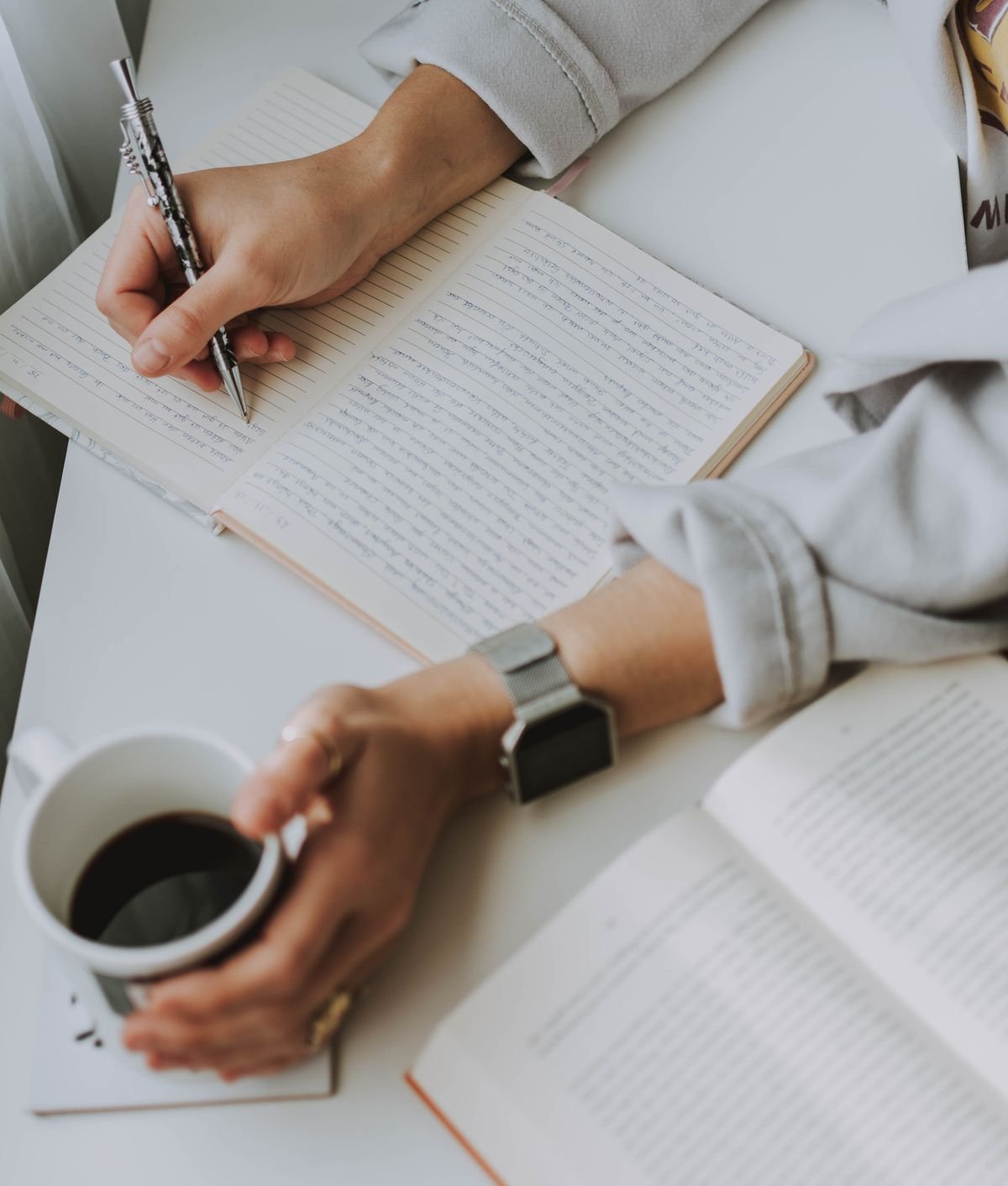 Person writing in a notebook while holding a coffee mug
